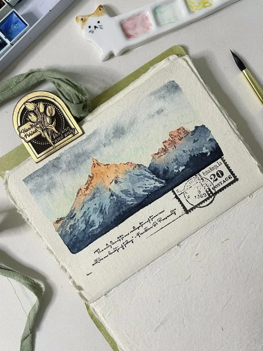 Watercolor tutorial image featuring a mountain landscape with snowy peaks, warm sunset lighting, and vintage postcard design elements.